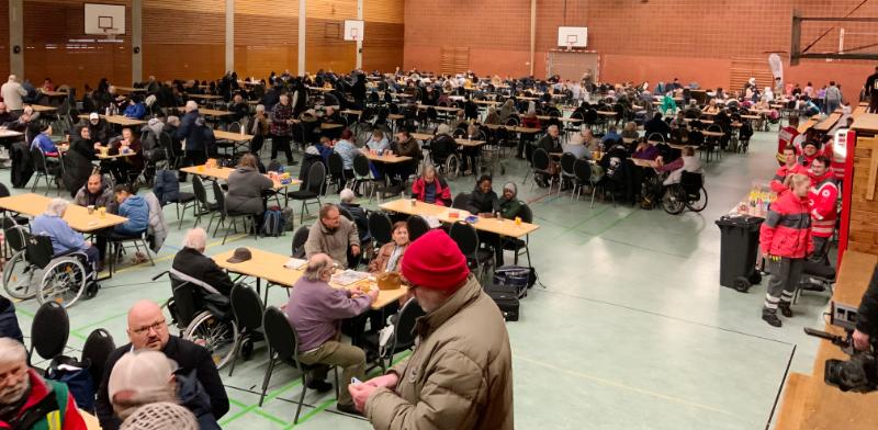 Menschen sitzen an Tischen in einer Turnhalle.