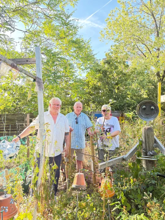 drei Menschen stehen fröhlich in einem grünen Naturgarten bei Sonnenschein und blauem Himmel.